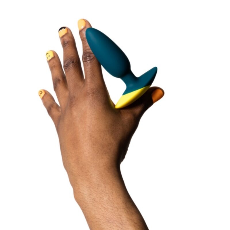 Hand with orange nail polish holding a teal silicone butt plug with a yellow base.
